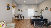 This single-floor community facility is thoughtfully designed to serve its occupants with a range of functional amenities. The reception area greets visitors with warm wood-look flooring, neutral gray walls, and recessed lighting. A dark-toned front desk provides a central workstation, while a bistro-height table with metal stools offers a casual gathering spot. Motivational wall art adds a welcoming, community-oriented atmosphere.

Adjacent to the reception area, a comfortable waiting room features wood-look flooring, plantation-shuttered glass doors that allow natural light to filter in, and seating along the wall. A pass-through window connects this space to a neighboring room, enhancing visibility and staff accessibility.

The fitness room is equipped with cardio machines including a treadmill, recumbent bike, and rowing machine, all set on a durable concrete floor. Gray walls, ceiling-mounted fluorescent lighting, and a window with blinds contribute to a clean and functional workout environment.

The laundry facility is expansive and efficiently arranged, with two long rows of commercial-grade washers and dryers lining each side of the room. The polished concrete floor and bright overhead lighting create a clean, practical space, with an exterior door at the far end providing convenient access to the outdoors.