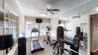 This fitness center is thoughtfully designed to support a well-rounded workout routine. The space is organized into distinct functional zones, beginning with a cardio area outfitted with treadmills and elliptical machines positioned along a windowed wall. Multiple windows fitted with horizontal blinds allow natural light to fill the room while offering views of an outdoor pool area, creating an energizing atmosphere during workouts. Wall-mounted televisions are conveniently placed for entertainment during cardio sessions.

The strength training area features a multi-functional cable machine and an adjustable weight bench, providing versatile options for resistance exercises. A dedicated free weights section includes a tiered dumbbell rack stocked with a range of weights, alongside a colorful medicine ball rack and kettlebells in various sizes, supporting functional fitness training.

Full-length mirrored panels line one wall, enhancing the sense of space and allowing users to monitor their form throughout their workout. The room is finished with neutral-toned carpeting, a textured ceiling, and ceiling fans that contribute to air circulation and comfort. French doors with glass panels connect this area to an adjacent space, adding an open, accessible quality to the overall layout.