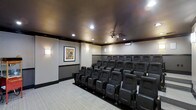 This single-floor private screening room delivers a true cinematic experience with a thoughtfully designed interior that balances function and elegance. Multiple rows of plush, dark leather recliner-style seats are arranged in a tiered, stadium-style layout, ensuring clear sightlines from every position in the room. A ceiling-mounted projector serves as the centerpiece of the entertainment setup, ready to deliver high-quality projection onto a dedicated screen. The room features a rich dark ceiling that enhances contrast and minimizes light reflection, contributing to an immersive viewing environment. Neutral-toned walls are accented by warm wall sconces that provide soft, ambient lighting along both sides of the room, creating a classic theater atmosphere. A framed piece of cinema-themed artwork adds a decorative touch that reinforces the room's identity. Light-toned carpeting runs throughout the floor, providing comfort underfoot and contributing to sound absorption. Subtle crown molding and dark trim work detail the transition between the walls and ceiling, adding a refined architectural finish. This space is purpose-built for private screenings, presentations, or entertainment gatherings, offering a polished and comfortable environment for any audience.