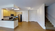 This two-floor space features an open-concept main living area that seamlessly integrates the kitchen and living room. The kitchen is equipped with light wood cabinetry, dark countertops, a tile backsplash, and stainless steel appliances, including a full-size refrigerator and microwave. A peninsula with a built-in sink adds both functionality and a natural division between the kitchen and the expansive living area. The flooring throughout is a warm-toned hardwood-style material that adds continuity and a welcoming feel. White walls and bright recessed and track lighting keep the space feeling airy and well-lit. Two interior white panel doors are visible from the main living area, providing access to additional rooms. The open layout of the main floor allows for flexible furniture arrangements, making the space highly versatile. The second floor adds additional square footage, expanding the overall living capacity of the unit. Clean architectural lines, neutral finishes, and a practical layout come together to create a functional and modern living environment suitable for a variety of lifestyles.