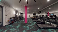 Located at 1081 N High St in Columbus, Ohio's vibrant Short North neighborhood, this contemporary fitness center is designed to deliver a high-energy workout experience. The space is defined by a striking aesthetic: bold pink structural columns rise from a richly patterned teal and black carpet, creating a dynamic visual contrast against crisp white geometric-patterned accent walls. Recessed lighting and ceiling fans are distributed throughout, ensuring the space stays well-lit and comfortable during any workout.

The cardio zone is generously equipped with a full row of True-brand treadmills, ellipticals, and stair climbers, each positioned for comfortable use. Large flat-screen TVs are mounted throughout the space, providing entertainment from virtually every workout station. Floor-to-ceiling mirrors along one wall amplify the sense of spaciousness and allow users to monitor their form.

A dedicated strength training area features a cable machine, weight bench, and a comprehensive rack of dumbbells to accommodate a wide range of training routines. A separate alcove houses additional strength machines, offering a quieter zone for targeted exercises. The overall layout is open and well-organized, providing ample room for movement and safe equipment use. The facility presents a polished, modern environment suited for a well-rounded fitness routine.