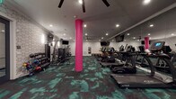 Located at 1081 N High St in Columbus, Ohio's vibrant Short North neighborhood, this contemporary fitness center is designed to deliver a high-energy workout experience. The space is defined by a striking aesthetic: bold pink structural columns rise from a richly patterned teal and black carpet, creating a dynamic visual contrast against crisp white geometric-patterned accent walls. Recessed lighting and ceiling fans are distributed throughout, ensuring the space stays well-lit and comfortable during any workout.

The cardio zone is generously equipped with a full row of True-brand treadmills, ellipticals, and stair climbers, each positioned for comfortable use. Large flat-screen TVs are mounted throughout the space, providing entertainment from virtually every workout station. Floor-to-ceiling mirrors along one wall amplify the sense of spaciousness and allow users to monitor their form.

A dedicated strength training area features a cable machine, weight bench, and a comprehensive rack of dumbbells to accommodate a wide range of training routines. A separate alcove houses additional strength machines, offering a quieter zone for targeted exercises. The overall layout is open and well-organized, providing ample room for movement and safe equipment use. The facility presents a polished, modern environment suited for a well-rounded fitness routine.