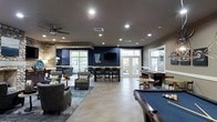 This single-floor community lounge is designed to offer a dynamic and inviting gathering space with a variety of entertainment and relaxation zones. The open-concept layout allows each area to flow seamlessly into the next, creating an expansive and social atmosphere throughout.

A prominent billiards table anchors one side of the room, positioned beneath recessed lighting that highlights the space's well-planned illumination scheme. Nearby, a stone fireplace adds a warm, rustic contrast to the room's otherwise modern aesthetic, flanked by comfortable lounge seating arranged over a patterned area rug.

Bold navy blue accent walls provide a strong design statement, complementing the neutral tile flooring laid in a herringbone pattern. A wall-mounted flat-screen television is positioned above a bar-height counter with seating, making it ideal for group viewing or casual socializing.

Large windows and French doors allow abundant natural light to fill the space while offering views of the surrounding exterior. A ceiling fan adds to the comfort of the lounge area. The combination of varied seating options, entertainment amenities, and thoughtful design details makes this a well-appointed communal space.