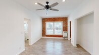Located at 1057 S Western Ave in Los Angeles, this single-floor unit blends classic character with modern updates throughout. The main living area makes an immediate impression with its striking exposed brick accent wall, which adds warmth and texture against the crisp white walls and ceilings. Wood-look flooring runs consistently throughout, complementing the bright, airy feel of the space. Recessed lighting and a contemporary ceiling fan with sleek blades ensure the room stays well-lit and comfortable year-round. Natural light filters in through a front-facing window framed by the exposed brick, offering views of the surrounding neighborhood. An open entryway area with clean white walls and light flooring provides a welcoming transition into the living space. The kitchen features white shaker-style cabinetry, a stainless steel refrigerator, and a window overlooking the exposed brick wall, creating a functional and visually cohesive cooking space. The bathroom is finished in an all-white palette, complete with a tub-shower combination, white tile surround, a window for natural ventilation, a single vanity with chrome fixtures, and a mirrored medicine cabinet. The overall layout is straightforward and efficient, with arched doorway details adding a subtle architectural touch throughout.