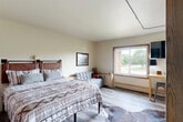 This efficiently designed single-floor space offers two bedrooms, a kitchen, and one bathroom within a thoughtfully arranged layout. The primary bedroom is generously proportioned, featuring light wood-style flooring, neutral walls, and a large sliding window that frames views of surrounding greenery, filling the room with natural light. Two closets — one a walk-in — provide ample storage, while a wall-mounted flat-screen TV, a compact workspace with bar seating, and a cozy sofa round out the room's functional and comfortable character. A cowhide-style area rug and framed wall art add a touch of character to the space. The kitchen is compact yet well-appointed, with rich wood cabinetry, a stainless steel range with a modern mosaic tile backsplash and a stainless steel hood vent, a sink with a dishwasher, and a large window overlooking an exterior brick facade that brings in additional natural light. Hardwood-style flooring continues throughout. The bathroom is designed with accessibility in mind, featuring grab bars near the toilet, a wide open layout with tile flooring, a large vanity with a granite-style countertop, a wide mirror with overhead lighting, and a separate toilet area for added privacy. The overall ambiance of the space is clean, practical, and welcoming.