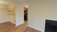 This single-story space features a straightforward, functional layout with a seamless connection between the main living area and the kitchen. The flooring throughout is a warm wood-look material, providing a cohesive and easy-to-maintain surface across the open floor plan. Walls are finished in a soft, neutral cream tone, creating a bright and versatile backdrop well-suited to a variety of interior styles.

A wide doorway opening connects the living space to the kitchen, which is equipped with dark wood cabinetry offering ample storage. Black appliances, including a range with overhead ventilation, are visible within the kitchen, complementing the darker cabinetry. A light-toned countertop is partially visible to the side, adding contrast to the kitchen's overall aesthetic.

White baseboards provide clean definition along the walls, contributing to a tidy, well-maintained appearance. Electrical outlets are conveniently placed along the walls, and a light switch is centrally located for ease of access. The open passage between rooms promotes good flow and makes the space feel connected and practical for everyday living.