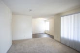 This single-floor unit offers a straightforward, functional layout with distinct living, sleeping, and kitchen zones. The living area is carpeted in neutral tones and features large vertical blinds covering a wide sliding door or window, allowing for adjustable natural light. An adjacent room with hardwood-style flooring connects through an open pass-through, offering flexibility in use.

The galley-style kitchen is lined on both sides with warm oak cabinetry providing ample storage, complemented by laminate countertops, a double-basin sink, and a full suite of black appliances including a range with overhead microwave and a refrigerator. Light hardwood-style flooring runs throughout the kitchen, adding warmth to the space.

The bedrooms are carpeted and bright, each featuring windows with horizontal blinds that allow natural light while maintaining privacy. The rooms offer generous proportions suitable for standard bedroom furnishings.

The bathroom includes a full tub and shower combination with clean white surround, a wood-toned vanity with countertop sink, and durable hardwood-style flooring. Neutral wall tones and consistent finishes throughout create a cohesive, understated aesthetic across the entire unit.