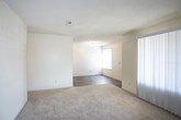 This single-floor unit offers a straightforward, functional layout with distinct living, sleeping, and kitchen zones. The living area is carpeted in neutral tones and features large vertical blinds covering a wide sliding door or window, allowing for adjustable natural light. An adjacent room with hardwood-style flooring connects through an open pass-through, offering flexibility in use.

The galley-style kitchen is lined on both sides with warm oak cabinetry providing ample storage, complemented by laminate countertops, a double-basin sink, and a full suite of black appliances including a range with overhead microwave and a refrigerator. Light hardwood-style flooring runs throughout the kitchen, adding warmth to the space.

The bedrooms are carpeted and bright, each featuring windows with horizontal blinds that allow natural light while maintaining privacy. The rooms offer generous proportions suitable for standard bedroom furnishings.

The bathroom includes a full tub and shower combination with clean white surround, a wood-toned vanity with countertop sink, and durable hardwood-style flooring. Neutral wall tones and consistent finishes throughout create a cohesive, understated aesthetic across the entire unit.