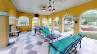 This spacious covered outdoor lounge area showcases a distinctive Mediterranean-inspired architectural design, characterized by a series of wide arched openings framed by pilasters set against vivid yellow stucco walls with contrasting light-colored banding. The open-air arches run along the perimeter, allowing abundant natural light and unobstructed views of the surrounding landscaped grounds, mature trees, and courtyard areas beyond a wrought iron fence line.

The flooring features a bold checkerboard pattern composed of large-format tiles in alternating light and dark tones, adding visual interest throughout the space. Overhead, a white ceiling is fitted with ceiling fans and decorative pendant lanterns that provide ambient lighting and air circulation.

The lounge is furnished with multiple seating arrangements, including teal-cushioned sofas, chairs, and wrought iron dining sets, creating distinct areas for relaxing and gathering. A built-in gas grill station situated near the perimeter adds functional outdoor cooking capability. Wall-mounted televisions are positioned for easy viewing from the seating areas. The overall ambiance is open, airy, and well-suited for community gatherings and leisure.