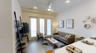 This single-floor space showcases a thoughtfully designed contemporary interior that balances comfort and style. The living area is anchored by a sleek, tufted gray sofa paired with a modern accent chair, centered around a geometric-patterned area rug in muted earth tones with warm orange accents. A white rectangular coffee table with a wire-frame base adds an industrial touch to the setting.

Natural light floods the room through a set of tall French doors that open onto a private balcony, offering views of the surrounding area. The doors are dressed with sheer white panels that gently diffuse the incoming light, creating a bright and airy atmosphere. A ceiling fan with a clean white finish and recessed lighting throughout ensure the space is well-lit at any hour.

The walls are finished in a crisp, neutral tone and are accented with framed artwork, contributing to the modern aesthetic. Light wood-toned flooring runs throughout, adding warmth and continuity to the space. A floor lamp in the corner and a lush indoor plant bring additional texture and life to the room.

The open layout allows for a seamless flow between living and dining areas, with the clean lines and minimal clutter reinforcing a sense of openness and intentional design throughout.