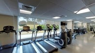 This fitness center is a thoughtfully designed single-floor facility built to support a comprehensive workout experience. The space is anchored by a row of modern cardio equipment, including multiple treadmills and elliptical machines, all positioned to take full advantage of the expansive floor-to-ceiling windows that line the perimeter. These large windows flood the room with natural light and frame views of lush tropical palm trees, creating an energizing and refreshing atmosphere for exercise. The clean, neutral color palette — with light-toned flooring, white walls, and a suspended ceiling with recessed panel lighting — contributes to a bright and open ambiance. A wall-mounted flat-screen television is positioned for visibility from the cardio equipment, adding an element of entertainment during workouts. The layout is well-organized, allowing for comfortable movement between machines. Ventilation is provided through ceiling-integrated HVAC units, ensuring a comfortable environment regardless of outdoor conditions. The overall design balances functionality with an inviting aesthetic, making this fitness center a polished and practical amenity suited for daily use.
