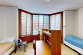 Located at 1825 R St NW in Washington, DC's vibrant Dupont Circle neighborhood, this single-floor room offers a functional and efficiently designed living space. The layout includes one bedroom, one bathroom, two closets, and an entrance area, all thoughtfully arranged to maximize usability.

The bedroom is the dominant feature of the space, characterized by a striking angled bay window that wraps around one corner of the room, allowing natural light to filter in through horizontal blinds while offering views of the surrounding brick building exterior. The ceiling features a drop-tile grid design, and the walls are constructed of painted concrete block, giving the space a solid, institutional character. Built-in warm wood cabinetry lines the base of the bay window, providing additional storage.

A central wood divider unit serves as both a room organizer and a desk partition, creating defined areas within the open bedroom. The light vinyl flooring runs throughout, keeping the overall aesthetic clean and uniform.

The entrance area provides a welcoming transition into the space, with access to two closets offering ample storage. The private bathroom features light blue square wall tiles surrounding a bathtub with shower curtain, a toilet, and a sink, delivering a clean and classic look. This self-contained room provides all essential amenities in a compact, well-structured layout.
