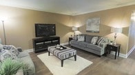 This single-floor space showcases a thoughtfully designed living area that blends comfort and contemporary style. The living room is finished with warm taupe walls accented by crisp white crown molding and baseboards, creating a clean, polished look throughout. Dark hardwood-style flooring grounds the space and contrasts beautifully with the plush, cream-colored area rug positioned at the center of the seating arrangement.

The room is furnished with a substantial grey upholstered sofa adorned with patterned decorative pillows, complemented by a striped ottoman that doubles as a central coffee table. A dark wood media console anchors one wall, supporting a large flat-screen television, while open shelving below provides practical storage. Ambient lighting is provided by a tall floor lamp, casting a warm, inviting glow across the room.

Wall art featuring a muted, abstract landscape adds a subtle artistic touch above the sofa. The overall layout is well-proportioned, offering a comfortable flow and a welcoming atmosphere suited for relaxing or entertaining. The cohesive color palette of grays, taupes, and blacks ties the entire space together with a modern yet approachable aesthetic.