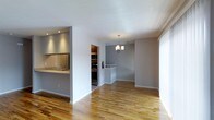 This two-floor space offers a well-designed, open-concept layout that seamlessly connects the kitchen and dining areas. The warm hardwood flooring extends throughout the main level, adding a welcoming character to the space. The kitchen features a raised countertop bar that serves as a natural divider between the cooking and living areas, complete with a sink, stainless steel appliances including an oven and microwave, and recessed lighting overhead. Adjacent to the kitchen, the dining area is defined by an elegant multi-arm chandelier that provides warm, focused lighting. The neutral gray and white wall palette creates a clean, modern aesthetic throughout. A staircase visible in the background connects to the second floor, accented by a white railing. Large windows allow natural light to fill the space, enhancing the bright and airy atmosphere. The overall design strikes a balance between functional and inviting, making it suitable for both everyday living and entertaining.