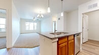 This well-appointed single-floor residence offers a thoughtfully designed open-concept layout that seamlessly connects the kitchen and living areas. The kitchen serves as the centerpiece of the space, showcasing warm knotty wood cabinetry, granite countertops, a large central island with a double sink and pendant lighting, and a full suite of stainless steel appliances including a refrigerator with ice maker, microwave, and range. Wood-look luxury vinyl plank flooring runs throughout the kitchen and entry areas, transitioning to plush neutral carpet in the living and bedroom spaces.

The living area is generously proportioned, bathed in natural light from large windows, and features both a ceiling fan and a decorative chandelier. A door provides direct access to an outdoor deck or patio area.

The bedrooms are comfortable and well-lit, each equipped with ceiling fans and windows that bring in natural daylight. The primary bedroom includes access to a spacious walk-in closet outfitted with multi-tier wire shelving for ample storage. A full bathroom features warm wood cabinetry with a marble-look countertop, a tub-shower combination, and easy-care wood-look vinyl plank flooring. The overall aesthetic is clean, neutral, and move-in ready.