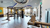 This well-appointed fitness center is organized across several distinct training zones, offering a comprehensive range of exercise options within a single, thoughtfully designed facility. The main cardio area is anchored by a row of treadmills, ellipticals, and stair climbers positioned along floor-to-ceiling windows that invite abundant natural light and outdoor views, creating an energizing workout environment. Large ceiling fans set within decorative circular coffers with blue LED accent lighting add a striking architectural element overhead.

The strength and functional training zone features a multi-station cable and power rack system, a suspended heavy bag, TRX suspension trainers, medicine balls, and a full dumbbell rack with accompanying benches, all arranged to maximize floor space. Full-length mirrors visually expand the area and support proper form during training.

A dedicated cycling room, accessible via sliding barn doors, houses multiple stationary and spin bikes alongside wall-mounted televisions. An adjoining functional training room offers a wall-mounted rig with resistance bands, suspension trainers, and additional heavy bag equipment on carpeted flooring. Throughout, bold yellow accent walls, recessed lighting, and durable flooring unify the space with a modern, high-energy aesthetic.