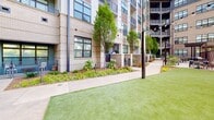 The outdoor amenity space at 3010 Edgeview Ln is a thoughtfully designed communal area set within a modern multi-story residential complex constructed with light brick and wood-panel accents. The grounds are organized into distinct functional zones connected by wide concrete walkways. A lush artificial turf lawn serves as the centerpiece, bordered by curated landscaping beds filled with native shrubs, ferns, ornamental grasses, and young trees that provide natural texture and greenery throughout the space. A decorative patterned brick wall adds a bold architectural accent, while black metal fencing encloses the perimeter for privacy and security. String lights are strung between dark metal poles across the turf area, creating an inviting ambiance for evening use. A central courtyard features contemporary outdoor lounge furniture arranged around a round fire pit table, ideal for social gatherings. Adjacent to this, a pergola-covered grilling and dining station is equipped with multiple gas grills and outdoor dining tables with seating, offering a dedicated space for outdoor cooking and entertaining. Individual townhome-style unit entrances line the walkway, each with private gated entries framed by landscaping. The overall design balances urban aesthetics with natural elements, creating a welcoming and functional outdoor retreat for residents.