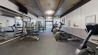 Located at 6300 Montgomery Blvd NE in Albuquerque, New Mexico, this fitness center offers a well-equipped exercise space designed to support a comprehensive workout routine. The room is characterized by a distinctive wood-paneled ceiling with exposed dark structural beams, ceiling fans providing ventilation, and recessed lighting that keeps the space bright and energized.

Large floor-to-ceiling windows along one wall flood the interior with natural light and offer views of the outdoor surroundings, including what appears to be a pool area and landscaped grounds. The flooring consists of durable rubber matting, appropriate for heavy equipment use and high-impact exercise.

The facility is outfitted with a wide variety of cardio equipment, including multiple elliptical trainers, treadmills, and stationary bikes, arranged to maximize the available floor space. A dedicated free weights area features a full dumbbell rack and adjustable weight benches, while a wall-length mirror visually expands the space and aids in form checks during workouts. A wall-mounted flat-screen television adds an entertainment element for users. Additional amenities include a water cooler, stability balls, and informational fitness charts posted on the walls. The overall ambiance is functional and well-maintained, making it a practical and inviting space for daily fitness activities.