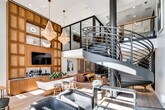 Located along Coast Highway in Dana Point, California, this sophisticated commercial space makes an immediate impression with its dramatic double-height ceilings and an eye-catching steel spiral staircase that serves as the architectural centerpiece of the interior. The staircase rises gracefully toward an open mezzanine level framed by sleek metal railings, adding both visual interest and vertical dimension to the space.

Floor-to-ceiling steel-framed windows along the street-facing façade flood the interior with abundant natural light, illuminating wide-plank light wood flooring throughout. A grand beaded chandelier anchors the main gathering area, complementing a full wall of warm, paneled wood cabinetry fitted with built-in shelving, a mounted flat-screen display, and a dark stone countertop with a sink — ideal for a reception or hospitality area.

The open layout accommodates multiple functional zones, including a lounge area furnished with leather seating, a central workspace with a solid wood desk and designer chairs, and an intimate meeting room enclosed by a vertical slat wood partition and dark-framed glass sliding doors. Coastal-inspired artwork and curated decor contribute to a refined yet welcoming ambiance throughout.