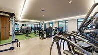Located at 57 Main St in Marlborough, MA, this fitness center offers a thoughtfully designed exercise room that maximizes its space with a comprehensive array of equipment and a clean, modern aesthetic. The room features a dual-zone flooring layout — a light concrete-style surface in the cardio and strength machine areas, and green turf flooring in the free weights zone — providing both functional variety and visual distinction throughout the space.

The cardio section is well-stocked with multiple elliptical trainers, treadmills, and stationary bikes, each equipped with individual screens for entertainment. A wall-mounted flat-screen TV adds a communal viewing option. The strength training area includes cable machines, power racks, barbells, and a full dumbbell rack, catering to a wide range of workout routines.

Large floor-to-ceiling windows and glass sliding doors line the perimeter, flooding the space with natural light and offering views of an outdoor courtyard and surrounding greenery. A full-length mirror in the free weights area enhances the sense of openness. Cove lighting along the tray ceiling adds a polished, upscale ambiance. The overall design balances functionality with a welcoming, contemporary atmosphere ideal for daily fitness use.