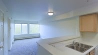 This single-floor space offers a clean, open-concept layout that integrates the kitchen and living area into one cohesive environment. The kitchen features a white countertop peninsula that serves as a natural divider, complete with a stainless steel sink — providing both functional workspace and a visual boundary between the two zones. The living area is finished with soft carpet flooring, contributing to a comfortable and quiet atmosphere. A flush ceiling light fixture provides overhead illumination, complementing the generous natural light that pours in through a pair of large windows. These windows also frame views of lush greenery outside, adding a natural element to the interior ambiance. The walls throughout are finished in crisp white, enhancing the sense of brightness and openness across the space. A baseboard heating unit runs along the exterior wall beneath the windows, ensuring efficient climate control. The overall aesthetic is minimal and functional, with a neutral palette that offers a versatile foundation for a variety of interior styles. The layout flows intuitively, making the most of the available space while maintaining an airy, uncluttered feel throughout.