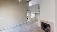 This two-story residence offers a well-organized layout across two floors, with the main living areas occupying the more spacious lower level and the bedroom suite positioned on the upper floor. The lower level centers on an open-concept living room distinguished by dramatically tall vaulted ceilings, neutral carpet, and a wood-burning fireplace framed in terracotta-toned tile, which serves as a prominent focal point. A sliding glass door provides access to a private balcony, introducing natural light into the living space. The kitchen is positioned adjacent to the living area and features white cabinetry, stainless steel appliances including a range, microwave, and refrigerator, a subway tile backsplash, and a granite-topped breakfast bar that opens toward the living room. A dedicated laundry area with a full-size washer and dryer is also found on this floor. The upper level houses the primary bedroom, a carpeted and generously proportioned room served by a ceiling fan and two closets. A full bathroom on this level includes a vanity with a framed mirror and a glass-enclosed shower. A second bathroom is also present within the unit. The interior throughout features neutral tones and carpet, contributing to a clean, cohesive aesthetic.