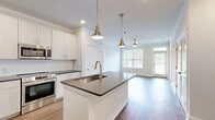 This thoughtfully designed 2-bedroom, 2-bathroom single-floor residence features a cohesive open-concept layout that seamlessly connects the kitchen, dining, and living areas. The modern kitchen serves as the centerpiece, boasting a large island with a dark quartz countertop, an undermount sink with a brushed gold faucet, white shaker-style cabinetry, a classic subway tile backsplash, and a full suite of stainless steel appliances including a gas range, microwave, and refrigerator. Brass pendant lights hang over the island, complementing the ceiling fan and natural light that pours in through large windows in the adjoining living area. Warm hardwood-style flooring runs throughout the entire residence, adding continuity and a welcoming feel. Crown molding accents the ceilings, lending a refined touch to every room. The spacious primary bedroom features large windows with outdoor views and a generous walk-in closet with wire shelving. Both bathrooms are finished with white vanity cabinetry, dark quartz countertops, undermount sinks, and brushed nickel hardware; the primary bath includes a glass-enclosed shower. A second well-sized bedroom and a hallway connecting all spaces complete this clean, contemporary home.