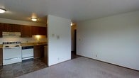 This single-floor unit offers a straightforward and functional layout designed to maximize everyday livability. The living area is finished with wall-to-wall carpeting, creating a warm and comfortable atmosphere, while crisp white walls provide a clean, neutral backdrop throughout.

Adjacent to the living space, the kitchen is efficiently arranged and features dark wood cabinetry that adds a touch of warmth and contrast to the light-toned walls. The kitchen is equipped with a sink, dishwasher, and under-cabinet lighting that illuminates the countertop workspace. The transition from the carpeted living area to the tile flooring of the kitchen clearly delineates the two spaces.

A partial wall separates the living area from the kitchen and hallway, offering a degree of definition between zones while maintaining an open, connected feel. A hallway leads further into the unit, suggesting additional private spaces beyond. Ceiling-mounted light fixtures provide consistent overhead lighting throughout, and a textured ceiling adds subtle visual detail to the interior. The overall ambiance is practical and move-in ready, suited for comfortable day-to-day living.