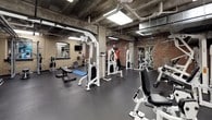 This fitness center offers a comprehensive workout environment spread across a single level, blending industrial architectural character with modern exercise equipment. The space is defined by its exposed brick walls, raw concrete ceilings with visible ductwork and piping, and structural columns that lend an authentic, loft-like aesthetic. Bold teal accent walls provide striking visual contrast throughout.

The cardio zone is equipped with multiple treadmills, elliptical machines, stair climbers, and stationary spin bikes, all positioned beneath recessed panel lighting and reflected in large wall mirrors that amplify the sense of space. A dedicated strength training area features a comprehensive selection of Hoist cable and selectorized machines, free-weight barbells, dumbbells, a weight bench, and a rack, catering to a full range of resistance training needs. An open functional training zone provides space for floor-based exercises, complete with stability balls, medicine balls, and a flat workout bench.

Rubber flooring throughout ensures durability and safety, while the overhead skylights and flush lighting maintain a bright, energizing atmosphere. The overall ambiance is motivating and purposeful, combining the raw charm of a repurposed industrial building with the practical layout of a fully functional fitness facility.