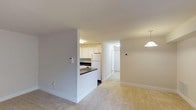 This single-floor residential space is designed with an open and functional layout that promotes easy flow between living areas. The kitchen is a standout feature, equipped with white cabinetry that extends to the upper walls, a dark contrasting countertop, and white appliances including a full-sized refrigerator. A partial wall with a raised breakfast bar separates the kitchen from the adjoining living and dining area, creating a sense of openness while maintaining a defined cooking zone. The flooring transitions from smooth, easy-to-clean surfaces in the kitchen to plush carpeting in the living area, adding warmth and comfort underfoot. Neutral-toned walls and white trim run throughout the space, lending a clean and cohesive aesthetic. A pendant light fixture adds a subtle decorative touch above the dining area. A hallway extends from the main living space, leading to additional rooms, with a white-paneled door providing privacy and access to the interior. The overall ambiance is bright, airy, and well-maintained, making the space feel welcoming and move-in ready. The straightforward design and neutral palette offer a versatile foundation for a variety of furnishing styles.