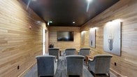 This single-floor media room offers a well-appointed space designed for comfortable viewing experiences. The interior is defined by warm, horizontal wood-paneled walls that lend a contemporary yet inviting character to the room. A dark, matte ceiling contrasts the natural tones of the walls, creating a visually enclosed atmosphere conducive to focused viewing.

A large wall-mounted flat-screen television serves as the room's focal point, positioned at a comfortable eye level for all seated guests. Recessed ceiling lighting provides soft, ambient illumination that complements the room's intimate ambiance. Wall-mounted sconces add additional accent lighting along the sides.

Seating consists of multiple plush, gray upholstered armchairs arranged in rows, each paired with small side tables for added convenience. The layout accommodates a modest group, making it well-suited for private screenings, presentations, or collaborative meetings.

Decorative touches, including framed artwork on the walls, add personality without disrupting the room's streamlined aesthetic. A whiteboard mounted to one wall suggests the space may also serve functional, collaborative purposes beyond entertainment.