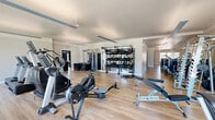 Located in Goodyear, Arizona, this fitness center at 2445 S Estrella Pkwy spans a generously sized single-floor layout divided into two dedicated exercise rooms, offering a comprehensive environment for a wide range of workout routines.

The main exercise room is expansive and bright, featuring light wood-look flooring, neutral wall tones, and large floor-to-ceiling sliding glass doors that open to views of an outdoor pool area, flooding the space with natural light. This room is outfitted with a robust selection of cardio equipment — including treadmills, ellipticals, stair climbers, and a Concept2 rowing machine — as well as an extensive array of strength training machines, free weights, adjustable benches, a TRX suspension training rig, and a full dumbbell rack. Floor-to-ceiling mirrors along one wall enhance the sense of space and allow for proper form monitoring.

The second exercise room serves as a dedicated cycling and studio space, equipped with ICG spin bikes and a ballet barre with mirrored wall, making it ideal for group fitness classes or individual low-impact training. Natural light enters through slatted window blinds, creating a clean and focused atmosphere.

A long, well-lit locker corridor connects the spaces, lined with rich wood-toned lockers and brightened by large windows. Together, the rooms provide a complete, modern fitness facility.