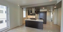 This single-floor space showcases a contemporary, open-concept layout centered around a well-appointed kitchen. The kitchen is a standout feature, designed with rich dark cabinetry that contrasts sharply against crisp white countertops on the central island, which serves as both a workspace and a visual anchor for the room. A stainless steel island range hood rises prominently above the cooking area, adding an industrial yet refined touch to the space. Full-height dark cabinetry lines the back wall, providing ample storage, while stainless steel appliances, including a refrigerator, integrate seamlessly into the design. The kitchen island includes a built-in cooktop, enhancing the functionality of the space. Light-toned hardwood flooring runs throughout, offering a warm counterbalance to the darker cabinetry and lending the space an airy feel. Recessed lighting and track lighting illuminate the kitchen effectively, while a window allows natural light to filter into the adjoining area. The open layout connects the kitchen to the living area, creating a cohesive and fluid living environment with a clean, modern aesthetic throughout.