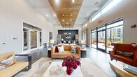 Located at 3 Lexington in Merrimack, New Hampshire, this single-floor community clubhouse showcases a sophisticated blend of contemporary design and warm, inviting elements. The expansive open-plan layout is anchored by soaring ceilings with a striking wood-clad tray ceiling detail, recessed lighting, and large floor-to-ceiling glass doors that flood the space with natural light and provide direct access to an outdoor terrace.

The main lounge area features large-format slate-toned tile flooring, complemented by curated seating including a caramel leather sofa, bold orange wingback chairs, and tufted banquette seating arranged around a sleek double-sided fireplace framed in dark subway tile and a natural wood surround. Abstract wall art and decorative accents add a refined, modern aesthetic throughout.

A fully equipped kitchenette area offers light wood cabinetry, a stainless steel French door refrigerator, a dark stone countertop with built-in sink, and a grey marble-style tile backsplash — ideal for hosting or casual gatherings. Patterned drapery panels, lush indoor plants, and abundant clerestory windows further enhance the bright, airy atmosphere, while views of the surrounding residential community reinforce the space's welcoming, communal character.