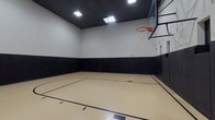 This indoor basketball court is a well-appointed athletic facility designed for serious play and safety. The court features a smooth, light-colored sport flooring surface with clearly marked boundary lines, a three-point arc, and a key, providing a regulation-style playing experience. A glass backboard with a standard orange rim and net is mounted on one end, offering professional-grade equipment for players of all skill levels.

The walls are finished in a two-tone design, with white upper walls rising to a considerable height and thick black padded panels lining the lower portion of all walls. These protective pads extend uniformly around the perimeter, minimizing injury risk during play and contributing to the court's clean, purposeful aesthetic. The dark ceiling contrasts with the white upper walls, adding visual definition to the space.

Lighting is provided by a combination of recessed ceiling fixtures and skylights, which allow natural light to supplement the artificial illumination, creating a bright and evenly lit environment. Multiple doors with emergency exit signage are integrated into the padded walls, ensuring safe and convenient access. Ventilation grilles are positioned throughout the upper walls, maintaining comfortable airflow. Overall, the court offers a functional, well-designed space suitable for basketball, fitness activities, and recreational use.