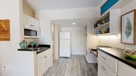 This efficiently designed single-floor studio makes smart use of its layout, seamlessly blending the living and kitchen areas into one cohesive space. The living area is anchored by a bold teal accent wall that adds a vibrant pop of color, complemented by light wood-look flooring that runs throughout. A ceiling fan overhead and a wall-mounted air conditioning unit keep the space comfortable, while French doors with blinds open to the outside, flooding the interior with natural light and providing easy outdoor access. A built-in shelving and desk unit lines one wall, offering dedicated workspace and display storage, topped with under-cabinet lighting for functional ambiance. The kitchen is thoughtfully integrated into the space, featuring white cabinetry, dark countertops, a stainless steel sink, an over-the-range microwave, and a full-size refrigerator. White subway tile walls add a clean, classic touch to the kitchen area. Open shelving provides additional storage and display space with under-shelf lighting. The bathroom is finished with tile walls and flooring, a vanity with mirror, and overhead cabinetry for storage. Throughout, the space balances practicality with a cheerful, modern aesthetic.