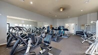 Located at 5150 N 99th Ave in Phoenix, Arizona, this fitness center offers a single, thoughtfully organized exercise room designed to support a full range of workout routines. The space features neutral gray walls, carpeted flooring, and recessed lighting complemented by a ceiling fan, creating a comfortable environment for exercise. Large arched windows along one wall invite abundant natural light and provide views of the surrounding outdoor area, enhancing the open and airy atmosphere of the room. A full-length wall mirror runs along one side, visually expanding the space and allowing users to monitor their form during workouts. The cardio section is well-stocked with multiple treadmills, elliptical trainers, stationary bikes, and a rowing machine, catering to a variety of aerobic exercise preferences. For strength training, the room includes cable and pulley machines, a lat pulldown station, and a dedicated free weights area featuring a tiered dumbbell rack and a flat bench. Wall-mounted televisions provide entertainment during workouts. A water vending machine is conveniently available on-site. Posted fitness center policies ensure a well-maintained and respectful environment for all users. This facility provides a comprehensive and accessible fitness option for residents of the Phoenix community.