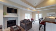 This single-floor space centers around a well-appointed living area designed for both comfort and functionality. The room's most notable architectural feature is the tray ceiling, which adds visual depth and a sense of elevated elegance to the space. Recessed lighting is embedded within the tray, providing soft, even illumination throughout.

The living area is anchored by a tiled fireplace with a clean, contemporary surround, paired with a wall-mounted flat-screen television above the mantle — a practical and streamlined arrangement. Plush carpeting covers the floor, contributing to the room's warm and inviting atmosphere.

Natural light fills the space through a front-facing window fitted with roller shades, while a set of French doors on the far wall opens to an outdoor area, offering additional brightness and easy exterior access. The neutral wall tones complement the overall palette, creating a cohesive and calming environment.

A dining area is partially visible to the right, suggesting an open or semi-open floor plan that connects living and dining functions seamlessly. Together, these elements make the space feel both practical and comfortable for everyday living.