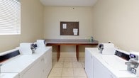 This shared laundry facility is a clean, functional, and thoughtfully arranged space designed to accommodate multiple users efficiently. The room is finished with neutral, warm-toned walls and light-colored tile flooring, creating a bright and welcoming environment.

At the heart of the space is a generous central folding counter with a laminate surface and wood-toned support legs, providing ample room for sorting and folding laundry. A cork bulletin board mounted above the counter serves as a convenient place for notices and information.

Lining the walls on both sides are multiple commercial-grade Speed Queen washers and dryers, offering reliable, high-capacity performance for heavy laundry loads. The machines are well-maintained and feature digital controls for ease of use. A utility sink is also available within the space for additional washing needs.

Adding to the convenience of the facility, a Coca-Cola vending machine is positioned near the entrance, providing refreshments during laundry sessions. A window fitted with horizontal blinds allows natural light to filter into the room. The space is accessed through a standard door marked with an illuminated exit sign, and a wall-mounted unit near the entrance suggests additional utility provisions. Overall, the laundry facility is practical, orderly, and well-suited for communal use.