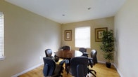 This single-floor professional space is centered around a well-appointed conference room designed for focused meetings and collaborative discussions. The room features a round wooden conference table surrounded by cushioned black leather executive chairs, providing comfortable seating for multiple attendees. The warm-toned beige walls create a calm, professional atmosphere, complemented by light hardwood flooring that adds a natural warmth to the space. Natural light filters in through a window fitted with horizontal blinds, allowing for adjustable illumination throughout the day. Recessed ceiling lights provide additional overhead lighting, ensuring the room remains well-lit regardless of the time of day. Decorative framed botanical prints adorn the walls, adding a refined aesthetic touch to the functional environment. A tall indoor plant in the corner introduces a refreshing natural element, enhancing the overall ambiance. Clean white baseboards and a smooth white ceiling complete the polished look of the room. This space is well-suited for small group meetings, client consultations, or team discussions, offering a balance of professional functionality and welcoming comfort in a compact, efficiently organized layout.