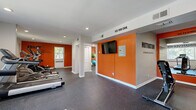 This fitness center is thoughtfully designed to maximize its single-floor layout, offering a comprehensive workout experience across three distinct zones. The main exercise area is immediately striking, featuring bold orange accent walls paired with neutral gray tones and large motivational quotes — including "YES YOU CAN," "DO NOT GIVE UP," and "MOTIVATION DETERMINES WHAT YOU DO" — that energize the space. Durable speckled rubber flooring covers the workout areas, providing both safety and easy maintenance. Recessed lighting throughout ensures a bright, well-lit environment.

The cardio zone is well-appointed with multiple Spirit treadmills and elliptical machines, complemented by natural light from windows along the walls. A secondary area houses a dumbbell rack with a full range of free weights, additional cardio equipment including a stationary bike, and a wall-mounted flat-screen television — ideal for both guided workouts and general use. Exercise balls and stretching equipment round out the available fitness tools.

An adjoining room provides extra space for functional training and equipment storage. The facility also includes a clean, classic bathroom featuring white square tile wainscoting, a soaking tub, and a window for natural ventilation — offering a convenient space to refresh after workouts.