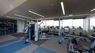 This contemporary fitness center is designed to deliver a premium workout experience within a clean, modern environment. The space is anchored by floor-to-ceiling windows that span an entire wall, flooding the interior with natural light and offering open views of the surrounding area — an energizing backdrop for any workout session. The ceiling features a dark, sophisticated finish contrasted by long, linear LED light fixtures that provide bright, even illumination throughout the space. The flooring consists of modular carpet tiles in a mix of blue and grey tones, adding a dynamic visual element while providing comfort underfoot. The facility is well-stocked with a diverse array of equipment, including multiple treadmills, a multi-function cable and weight machine, a rowing machine, and an assortment of free weights and benches. Cardio machines are thoughtfully positioned facing the windows, allowing users to take in the views while training. A mirrored wall on one side enhances the sense of openness and supports proper form during strength exercises. The overall ambiance is professional and motivating, combining thoughtful layout with high-quality equipment to create a fully functional, well-appointed fitness facility.