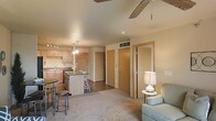 This single-floor residence offers an open-concept layout that seamlessly connects the kitchen and living area, creating a cohesive and functional living space. The kitchen is equipped with light wood cabinetry, a dark-toned countertop island with bar-height seating, a sink along the perimeter wall, and track lighting overhead — providing ample workspace and casual dining options. Pendant lighting above the island adds a subtle decorative touch.

The living area is finished with wall-to-wall carpeting in a neutral beige tone, contributing to a warm and comfortable ambiance. A ceiling fan with wooden blades is centrally positioned, supporting air circulation throughout the space. The room receives ambient interior lighting and is furnished with a light blue upholstered chair accompanied by a small side table and lamp.

Toward the back of the unit, several interior doors are visible — suggesting access to a bedroom, closet, and bathroom — all framed with light wood trim that complements the kitchen cabinetry. The consistent use of warm neutral tones across walls, flooring, and woodwork gives the space a unified and understated aesthetic throughout.