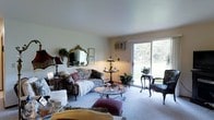 Located at 3220-3224 Mary St in Marinette, Wisconsin, this single-floor property offers a comfortable and welcoming layout well-suited for everyday living. The living room serves as the heart of the space, featuring neutral walls and light carpeting that create a warm, inviting atmosphere. A large sliding glass door floods the room with natural light and provides direct views of the lush, tree-lined outdoor surroundings, seamlessly connecting the interior to the outdoors. The room comfortably accommodates a classic seating arrangement, including a floral-patterned sofa, upholstered armchairs, and a glass-topped coffee table, all centered around an ornate framed mirror that adds a touch of traditional elegance to the décor. A wall-mounted air conditioning unit ensures year-round comfort. Decorative plants and table lamps are thoughtfully placed throughout, enhancing the homey character of the space. The overall ambiance is relaxed and charming, reflecting a traditional aesthetic with practical amenities, making this Marinette property a comfortable single-level retreat.
