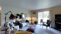 Located at 3220-3224 Mary St in Marinette, Wisconsin, this single-floor property offers a comfortable and welcoming layout well-suited for everyday living. The living room serves as the heart of the space, featuring neutral walls and light carpeting that create a warm, inviting atmosphere. A large sliding glass door floods the room with natural light and provides direct views of the lush, tree-lined outdoor surroundings, seamlessly connecting the interior to the outdoors. The room comfortably accommodates a classic seating arrangement, including a floral-patterned sofa, upholstered armchairs, and a glass-topped coffee table, all centered around an ornate framed mirror that adds a touch of traditional elegance to the décor. A wall-mounted air conditioning unit ensures year-round comfort. Decorative plants and table lamps are thoughtfully placed throughout, enhancing the homey character of the space. The overall ambiance is relaxed and charming, reflecting a traditional aesthetic with practical amenities, making this Marinette property a comfortable single-level retreat.
