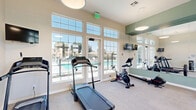 This fitness center in Moreno Valley, CA offers a thoughtfully designed, single-floor exercise space that provides a comprehensive workout environment. The room features neutral-toned walls, light carpeted flooring, and high ceilings that lend a sense of openness and comfort. An abundance of natural light pours in through large, grid-pane windows and glass-panel doors, with views of an outdoor pool area and surrounding landscape — creating an inviting and energizing atmosphere for exercise.

The space is outfitted with a well-rounded selection of fitness equipment. Cardio offerings include multiple treadmills and stationary cycling bikes, some equipped with digital displays for interactive training. A dedicated cycling area along one wall features spin bikes and motivational framed artwork, adding personality to the space. Strength training is supported by a barbell bench press station, a weight rack with free weights, and additional resistance machines.

A full-length wall mirror with a sage green accent border visually expands the space and aids exercisers in monitoring their form. Wall-mounted flat-screen televisions are positioned for convenient viewing during cardio sessions. Overhead pendant lighting complements the natural light, ensuring the space remains well-lit at all hours. A glass-paned interior door connects to an adjacent area, adding convenience and accessibility to the overall facility layout.