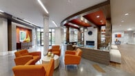 Located at 322 Baldwin Rd in Odenton, Maryland, this expansive single-floor community clubhouse offers a sophisticated and welcoming environment designed for both socializing and productivity. The space makes a strong first impression with its grand, open lobby, featuring polished stone tile flooring, bold dark wood columns, and a sweeping curved ceiling detail accented in a warm orange-red tone that frames a striking stone fireplace. Vibrant orange armchairs and contemporary pendant lighting anchor the central lounge, creating an inviting focal point. Adjacent areas transition seamlessly from tile to patterned carpet, defining distinct zones throughout the open-plan layout. A dedicated workstation area provides built-in desks and task seating, while glassed-in private offices and a bar-height counter workspace offer additional productivity options. A comfortable lounge zone features plush sofas, a wall-mounted TV, warm wood cabinetry, and a coffee bar. Casual seating nooks with tufted benches and sculptural pendant lights line the corridors. Floor-to-ceiling windows throughout flood the space with natural light and frame views of the surrounding greenery. The overall ambiance blends modern design with warm, residential comfort, making this a well-appointed and functional community gathering space.