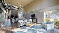 This single-floor community clubhouse, identified as Campbell Run, offers an impressive blend of industrial and rustic design elements unified under dramatically vaulted ceilings with exposed wooden trusses. The expansive main gathering area serves as the centerpiece of the space, featuring a striking floor-to-ceiling brick fireplace, warm wood-look flooring, and an abundance of natural light streaming through oversized windows and tall glass entry doors that frame views of the surrounding landscape.

Corrugated metal wall paneling and visible HVAC ductwork lend an intentional industrial character throughout, balanced by soft, neutral furnishings and warm pendant and sconce lighting. Multiple seating zones are thoughtfully arranged across the open floor plan, including a lounge area anchored by a sofa, accent chairs, and a rustic wood coffee table, as well as several intimate conversation clusters ideal for social gatherings.

A dining area with a dark wood farmhouse-style table and bench seating sits adjacent to decorative open metal shelving against an exposed brick wall, providing both function and visual interest. Sliding barn-style doors and louvered partitions define zones while maintaining the open, airy atmosphere. The overall ambiance is welcoming and modern-rustic, designed to encourage community interaction and relaxed gathering.
