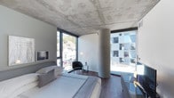 This single-floor space showcases a striking blend of industrial and contemporary design, defined by its exposed raw concrete ceiling and sleek architectural detailing. A prominent cylindrical concrete column anchors the room, adding sculptural character to the open layout. Floor-to-ceiling windows wrap around the space, flooding the interior with natural light and offering expansive views of the surrounding urban environment, including glimpses of neighboring modern architecture and a tree-lined hillside.

The bedroom area features a neatly appointed low-profile bed dressed in neutral linens, complemented by dark hardwood flooring that adds warmth against the cool concrete overhead. A slim, wall-mounted television is positioned opposite the bed for convenience. Thoughtfully placed accent pieces include a rounded lounge chair, a minimalist side table, and a slender floor lamp that adds ambient illumination. The overall furnishings are understated and curated, maintaining a calm, sophisticated palette of grays and whites.

The abundant natural light, open sightlines, and interplay of raw and refined materials create an ambiance that is both refined and edgy — ideal for those who appreciate modern architectural design with an urban sensibility.