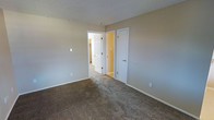 This single-floor residential space offers a clean and functional layout designed for comfortable everyday living. The interior is finished with neutral beige-toned walls that create a calm, understated ambiance throughout. The flooring features wall-to-wall carpet in a warm brown tone, adding softness and a sense of coziness underfoot.

The room visible in the space is generously proportioned, providing ample room for furnishings and movement. A textured popcorn ceiling adds a classic residential character overhead. Two white paneled doors are prominently featured — one leading to an adjacent hallway or common area, illuminated by warm yellow-toned lighting, and a second door that likely accesses a closet, offering convenient built-in storage. White baseboards frame the perimeter of the room, providing a crisp contrast against the neutral walls and carpet.

The overall layout suggests a well-connected interior, with the hallway beyond offering access to additional areas of the home. The space is bright, open, and move-in ready, presenting a straightforward and practical residential environment suited for a variety of uses and furnishing styles.